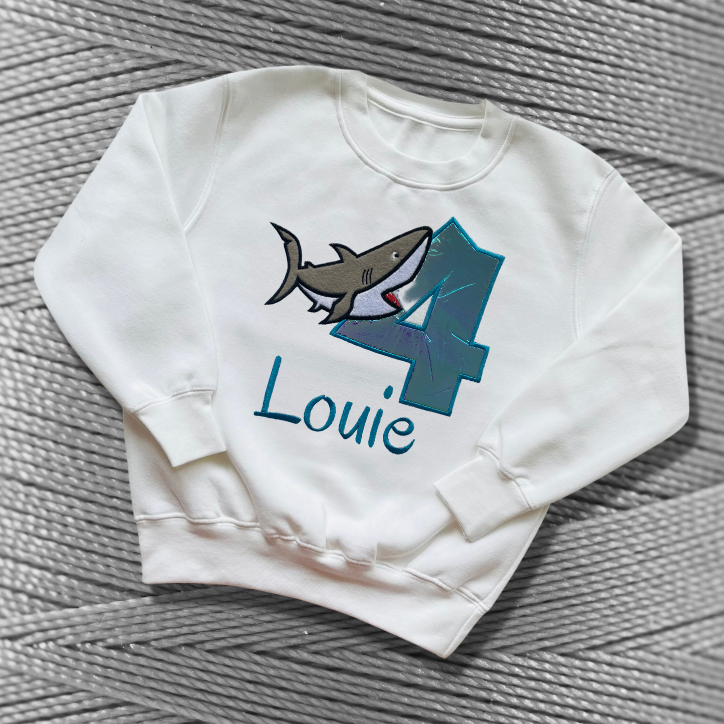 Childrens white sweatshirt with fleece lining. Embroidered applique on the front with the birthday age, shark design and the childs name.