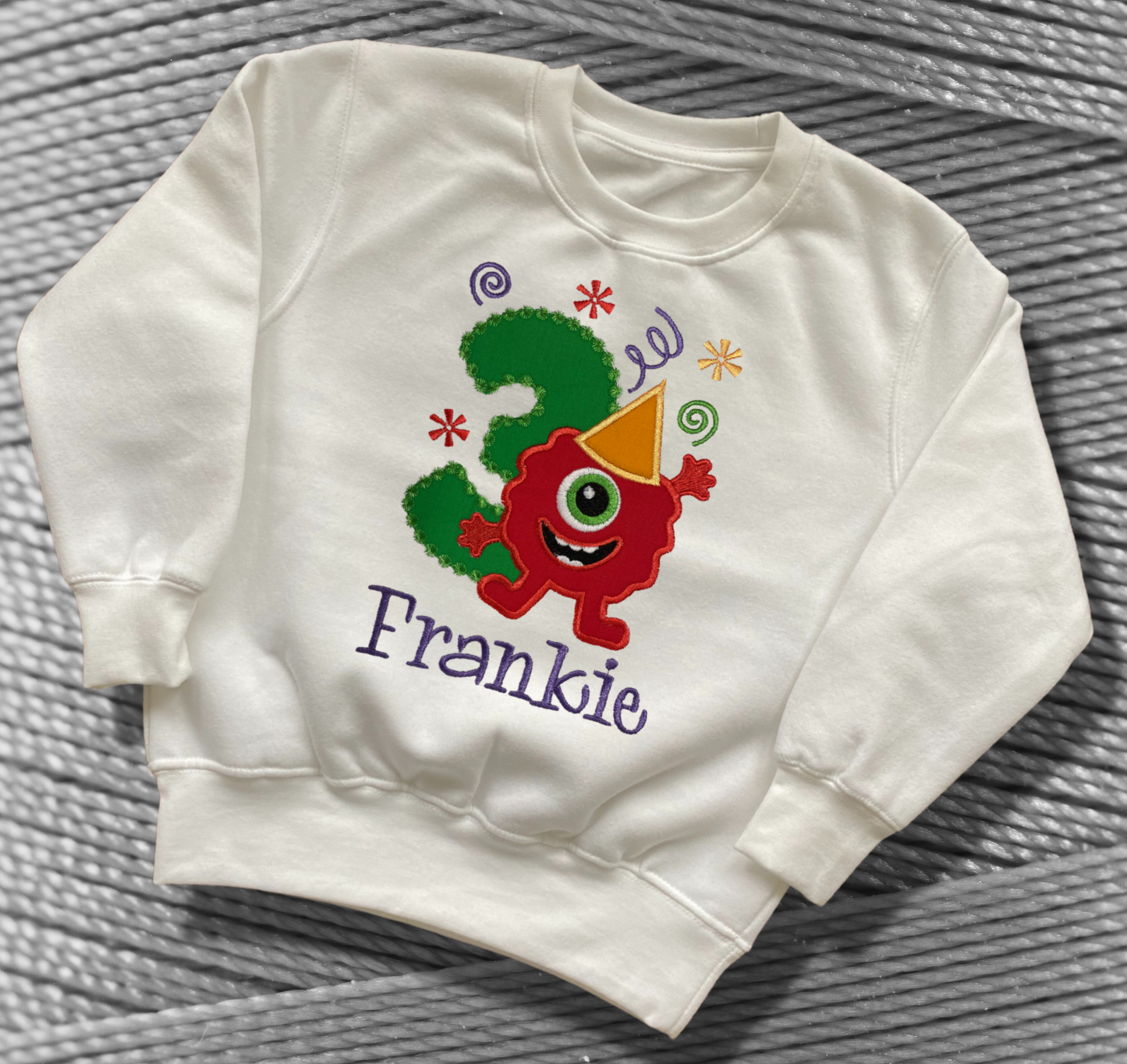 Childrens white sweatshirt with fleece lining. Embroidered applique on the front with the birthday age, funny monster design and the childs name.