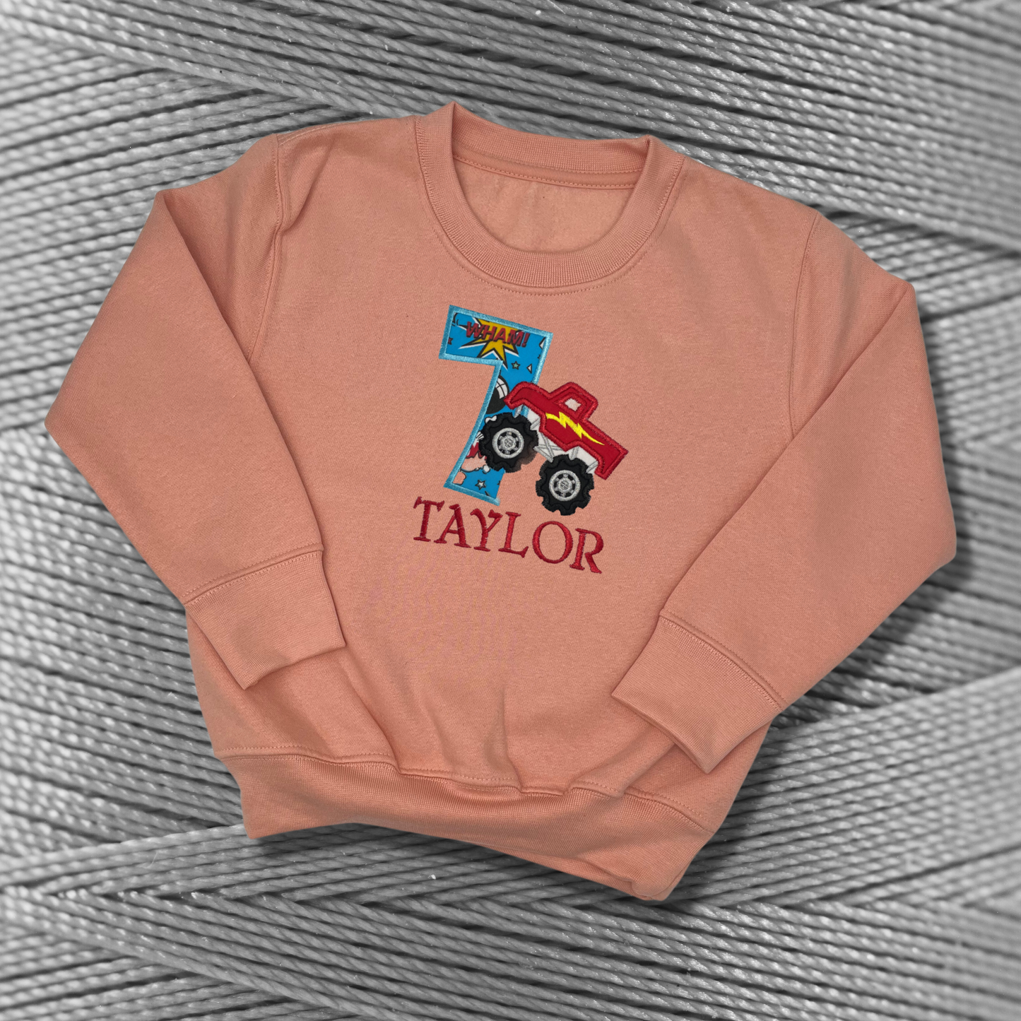 Childrens peachy-pink sweatshirt with fleece lining. Embroidered applique on the front with the birthday age, monster truck design and the childs name.