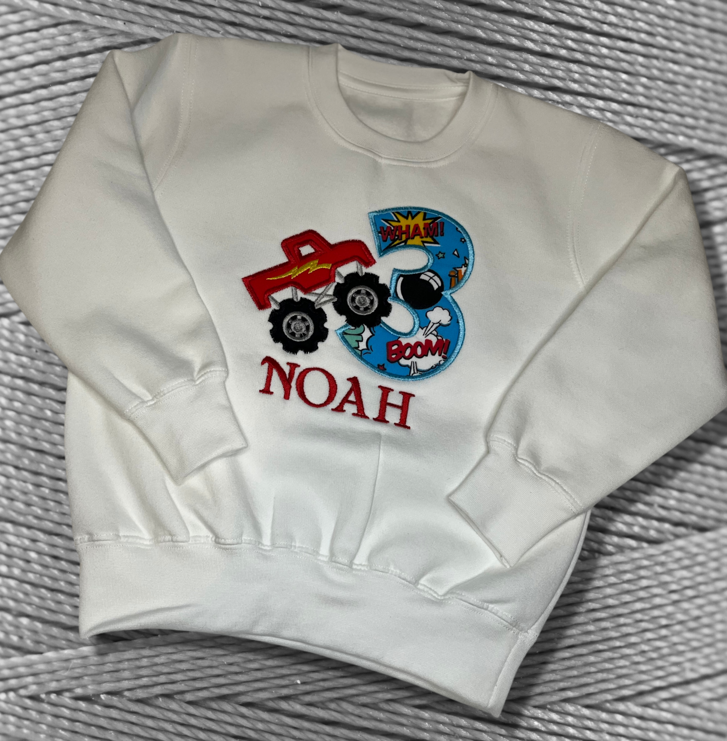 Childrens white sweatshirt with fleece lining. Embroidered applique on the front with the birthday age, monster truck design and the childs name.