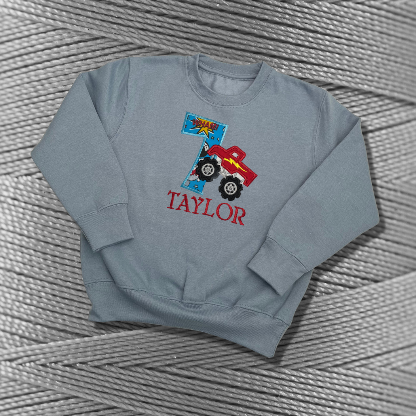 Childrens light blue sweatshirt with fleece lining. Embroidered applique on the front with the birthday age, monster truck design and the childs name.