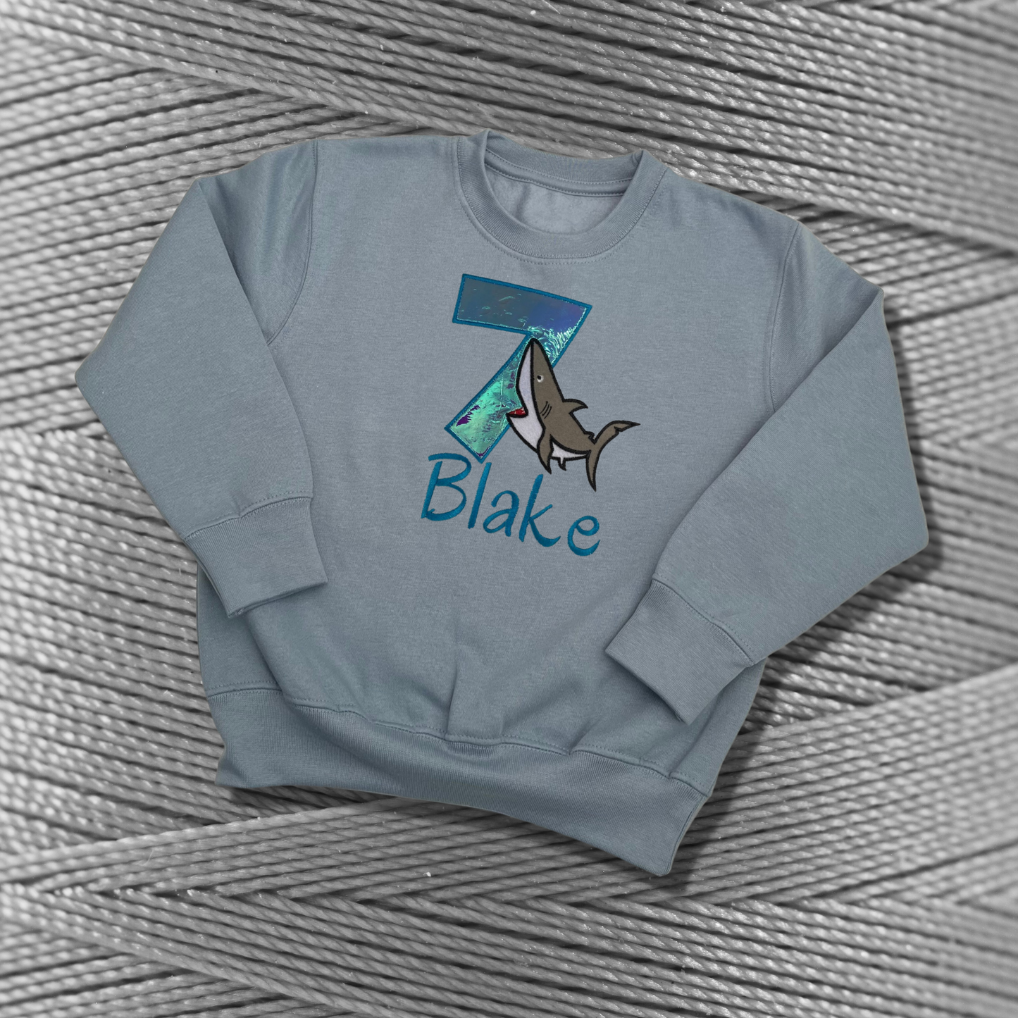 Childrens light blue sweatshirt with fleece lining. Embroidered applique on the front with the birthday age, shark design and the childs name.