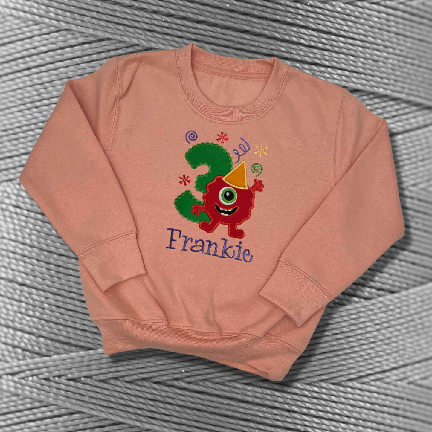 Childrens peachy-pink sweatshirt with fleece lining. Embroidered applique on the front with the birthday age, funny monster design and the childs name.