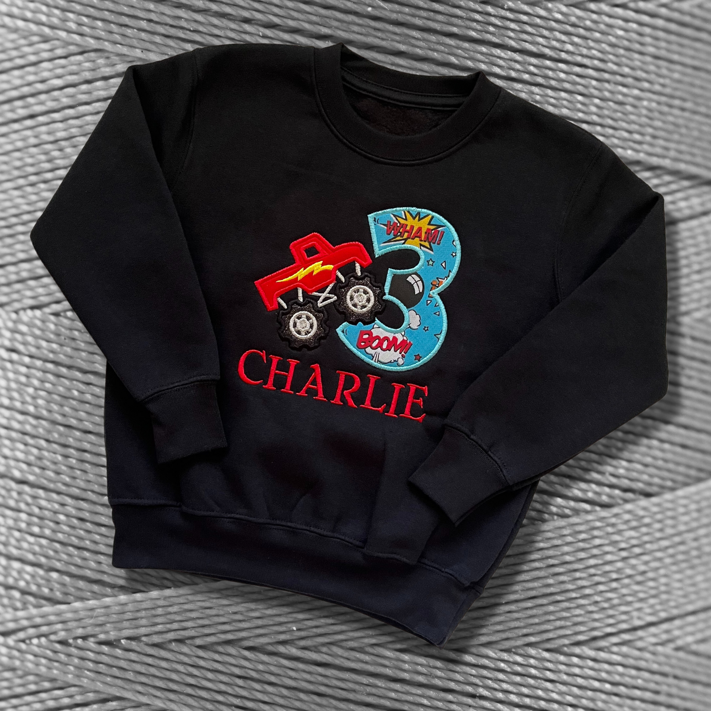 Childrens black sweatshirt with fleece lining. Embroidered applique on the front with the birthday age, monster truck design and the childs name.