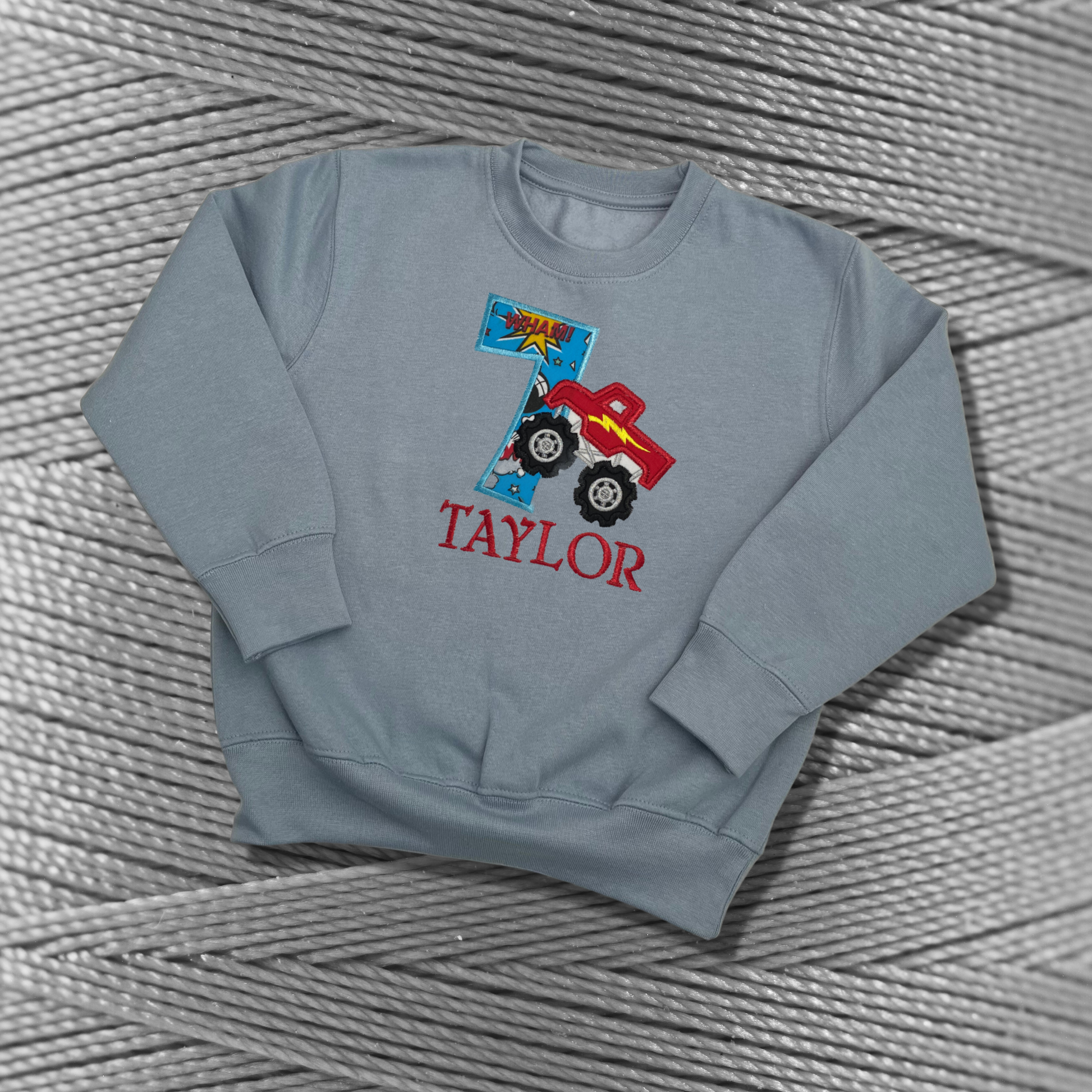 Childrens light blue sweatshirt with fleece lining. Embroidered applique on the front with the birthday age, monster truck design and the childs name.