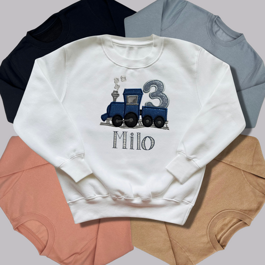 Blue train design embroidered on a white sweatshirt with other coloured sweatshirts behind